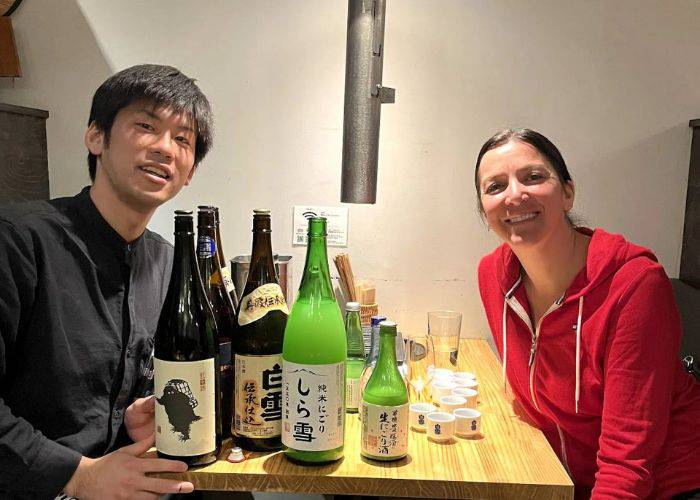 A guest and an expert sake guide sitting and smiling at the camera. On the table, sake bottles and cups.